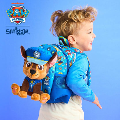 A young child wearing a blue Paw Patrol backpack with a plush Chase character attached to the back, posed against a bright blue and pink studio background. The Paw Patrol and Snuggle logos are visible in the upper left.